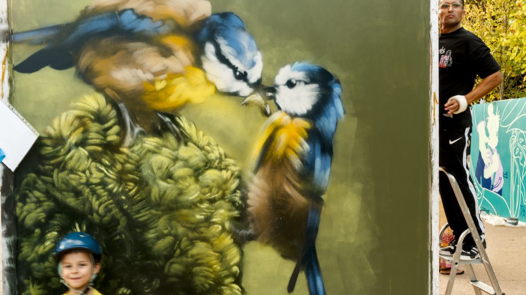 Un enfant, pull jaune et casque de cycliste bleu, pose devant une peintre de street art représentant une mésange nourrissant son petit, dans les mêmes couleurs. Au fond, on voit un artiste sur son escabeau. © Jean-Claude Marguerite 